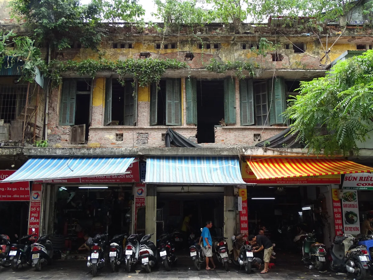 A weathered old colonial building with shuttered windows towers above street-level shops and parked motorbikes in Hanoi's.