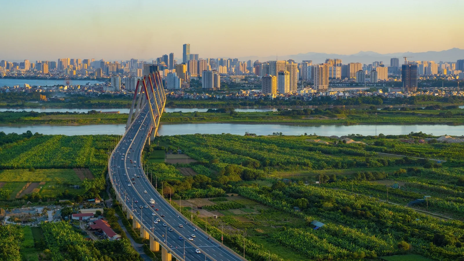 A cable-stayed bridge curves over the Red River toward Hanoi's skyline, flanked by green farmland at sunset.