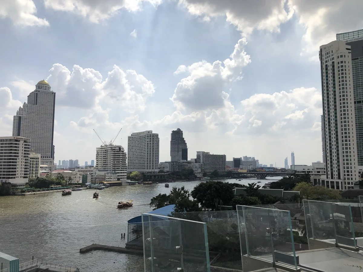 High-rise towers line the Chao Phraya River in Bangkok under a cloudy sky, with boats crossing the water below.