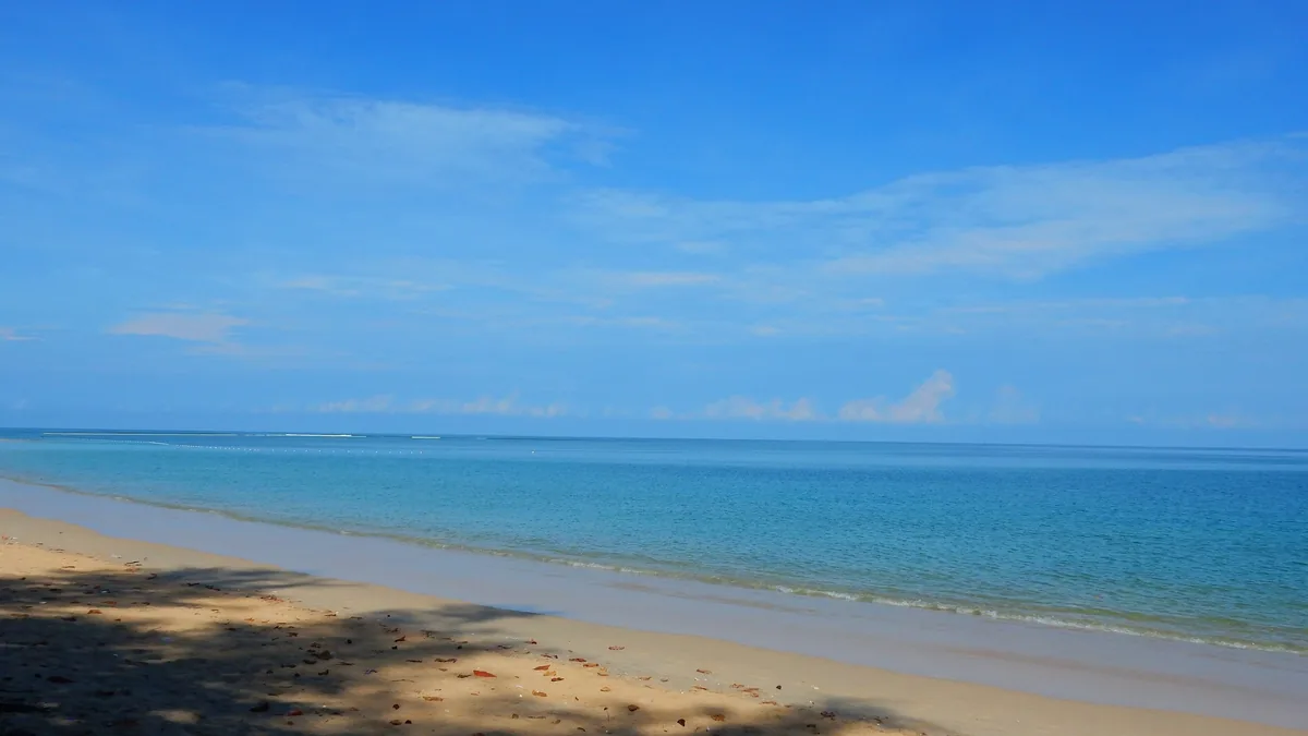 A quiet sandy beach meets calm turquoise water under a wide blue sky in Phuket.