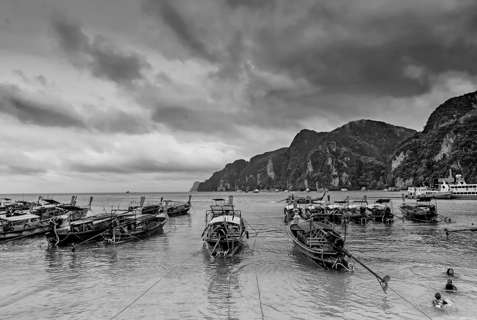 Longtail boats moored in shallow water beneath dramatic clouds and limestone cliffs near Krabi, Thailand.