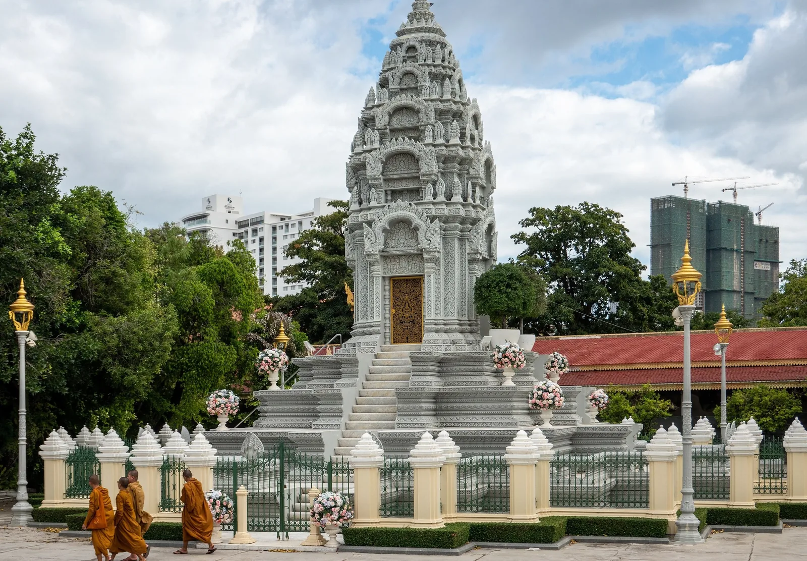 Monks in orange robes walk past a white stupa-shaped stone memorial with city towers in the background in Phnom Penh.