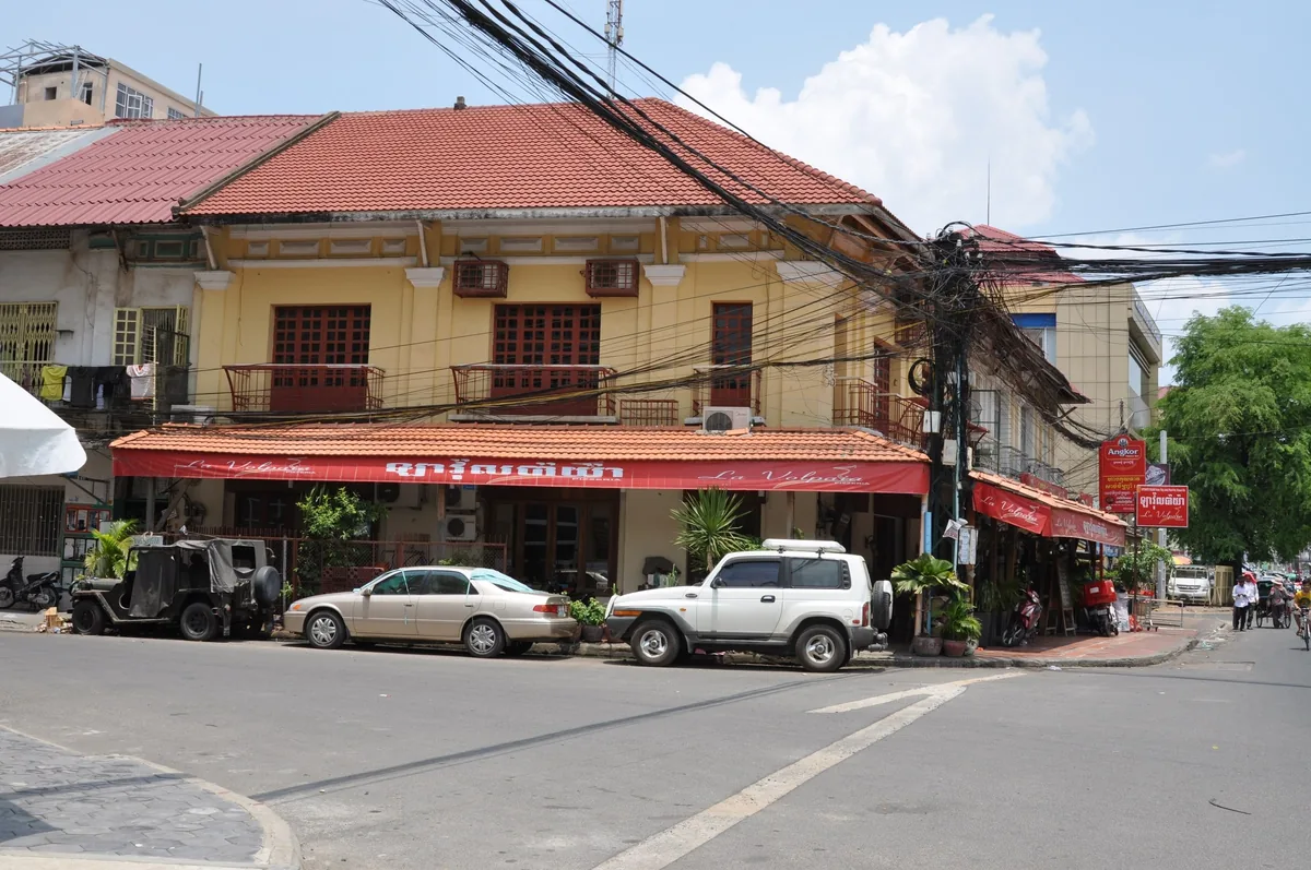 A colonial-era yellow building with red tiled roof and shophouses lines a street corner in Phnom Penh, with cars parked.
