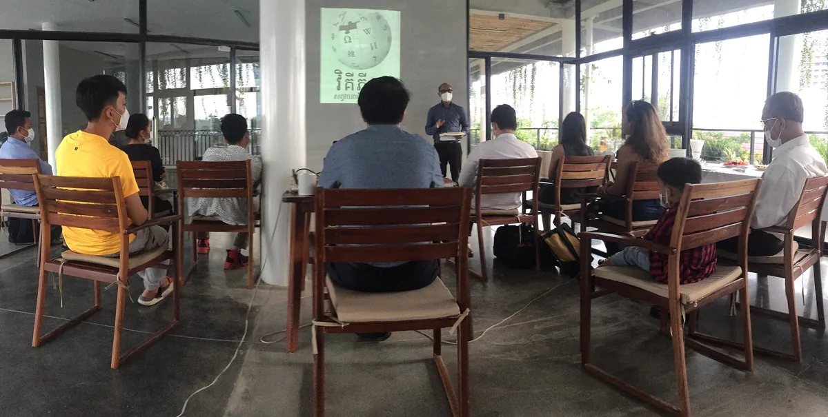A small group attends a presentation with a projected logo in a bright, open room in Phnom Penh.