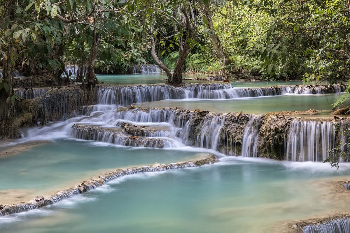 Tiered turquoise pools cascade over limestone terraces surrounded by lush forest at Kuang Si Falls near Luang Prabang.