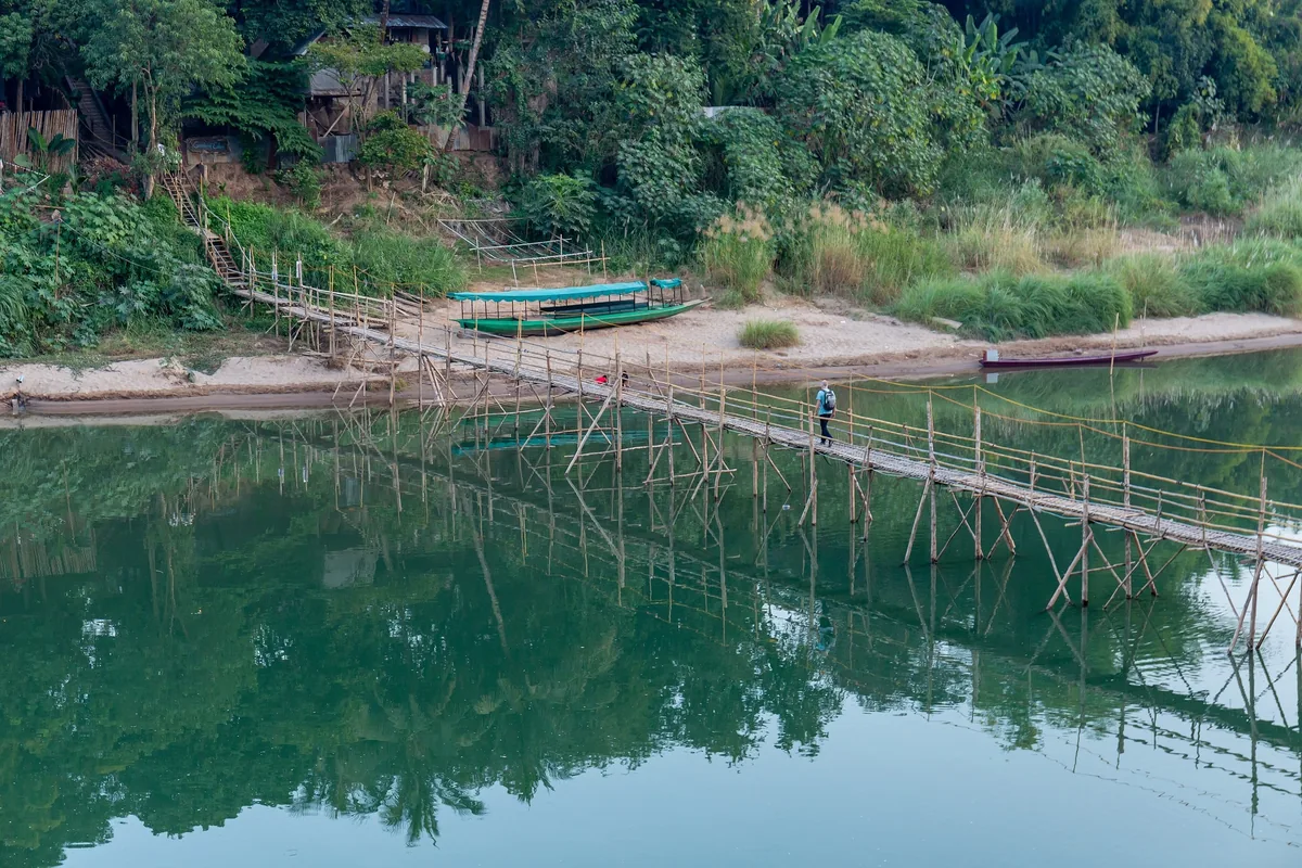 A person crosses a bamboo bridge over the Nam Khan River near Luang Prabang, with wooden boats moored on the sandy bank.