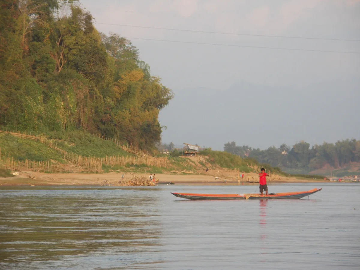 A man in a red shirt stands on a wooden boat on the Mekong River near Luang Prabang.