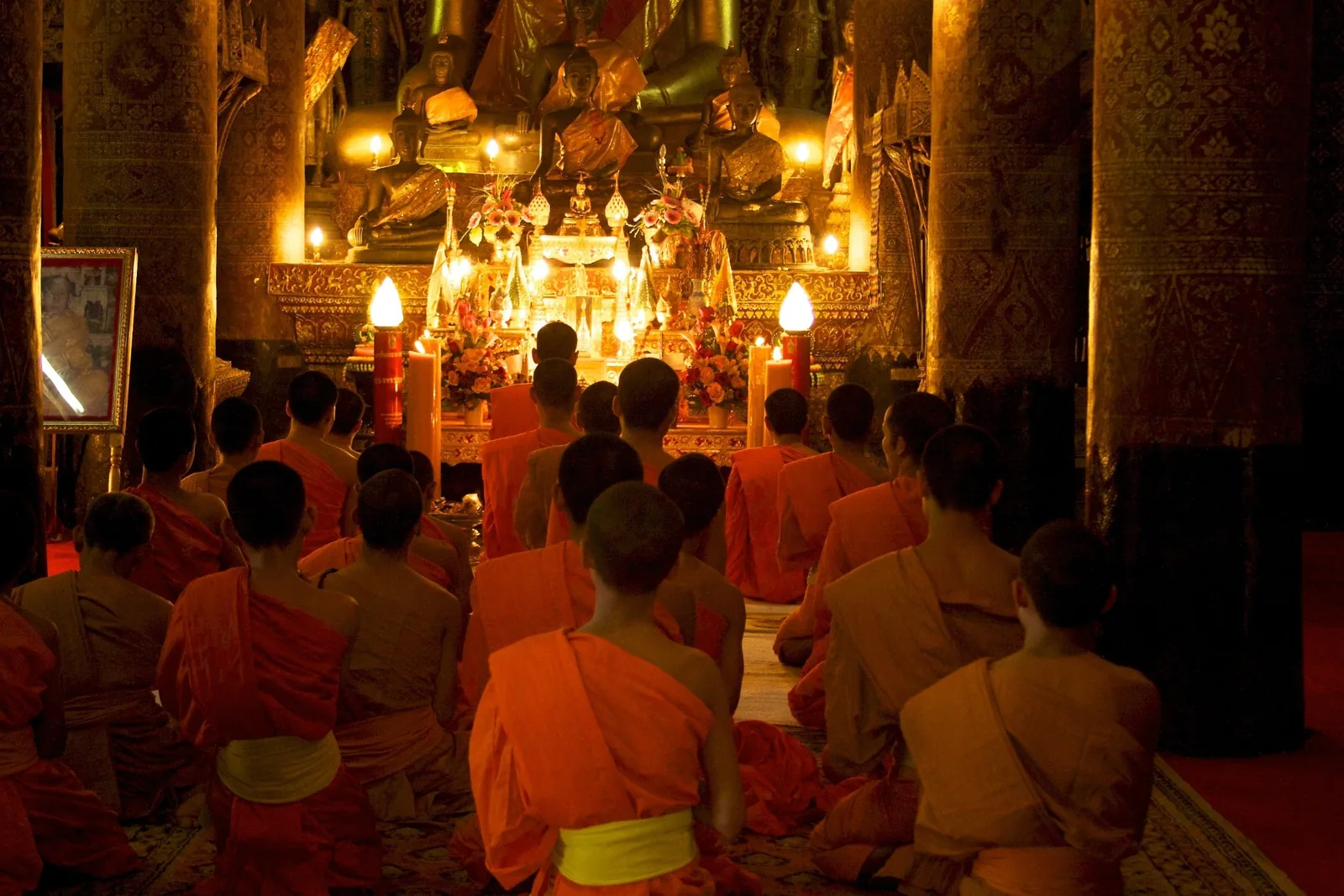 Young monks in orange robes kneel in prayer before a gilded Buddha altar inside a temple in Luang Prabang.