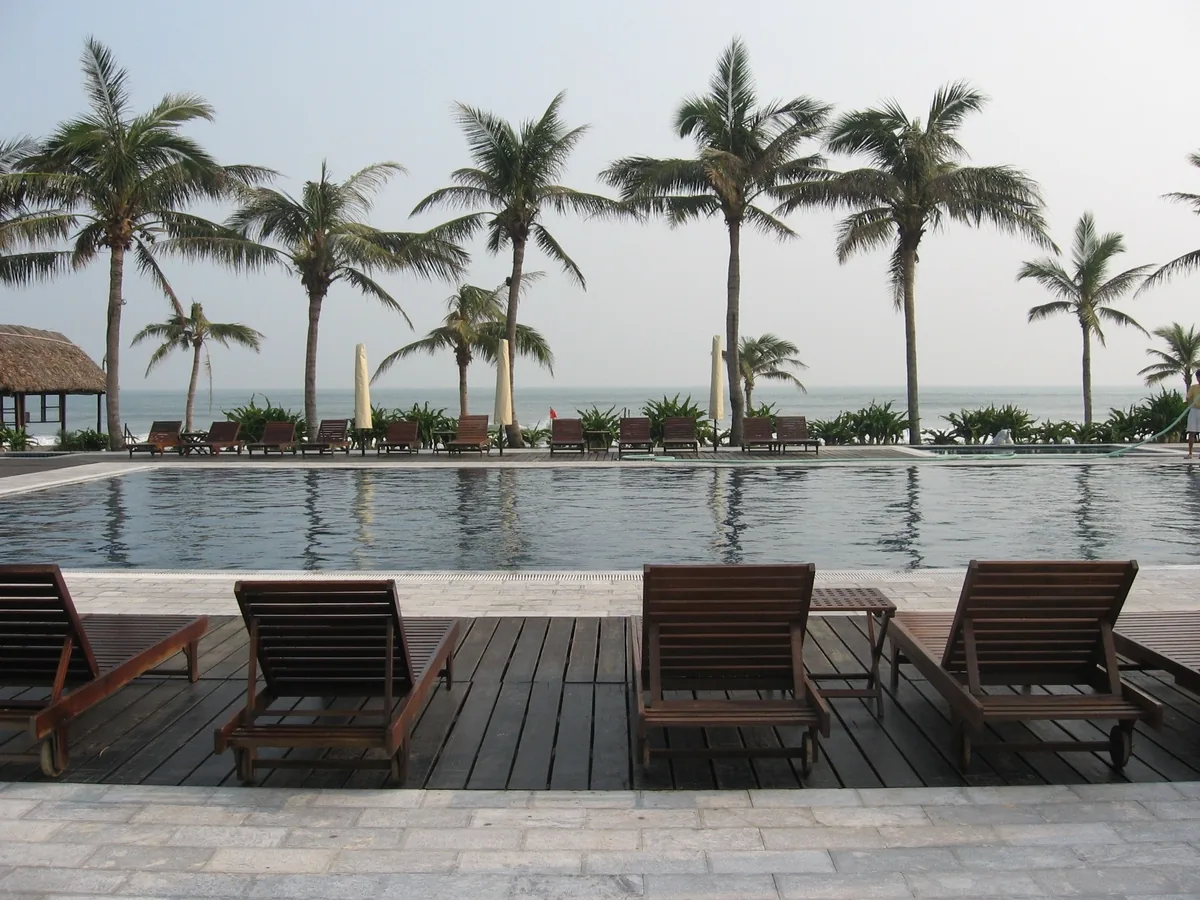 Wooden loungers line a resort swimming pool framed by palm trees near the beach in Hoi An.