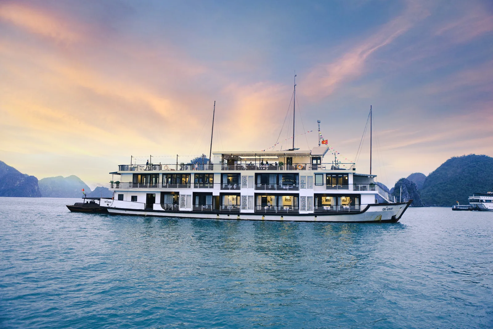 A multi-deck cruise ship anchored among limestone karsts at sunset in Halong Bay.