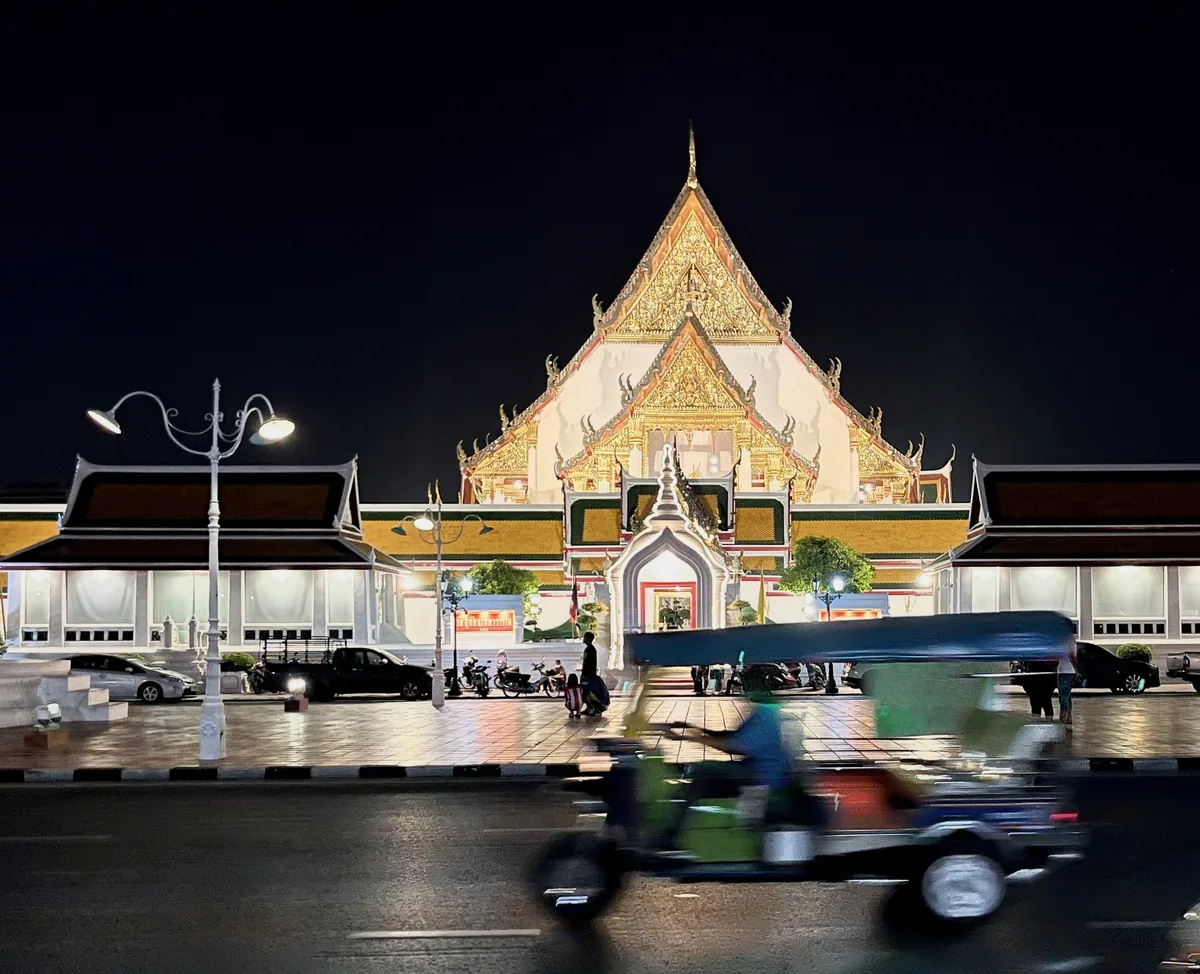 A tuk-tuk speeds past illuminated Wat Suthat temple in Bangkok at night.