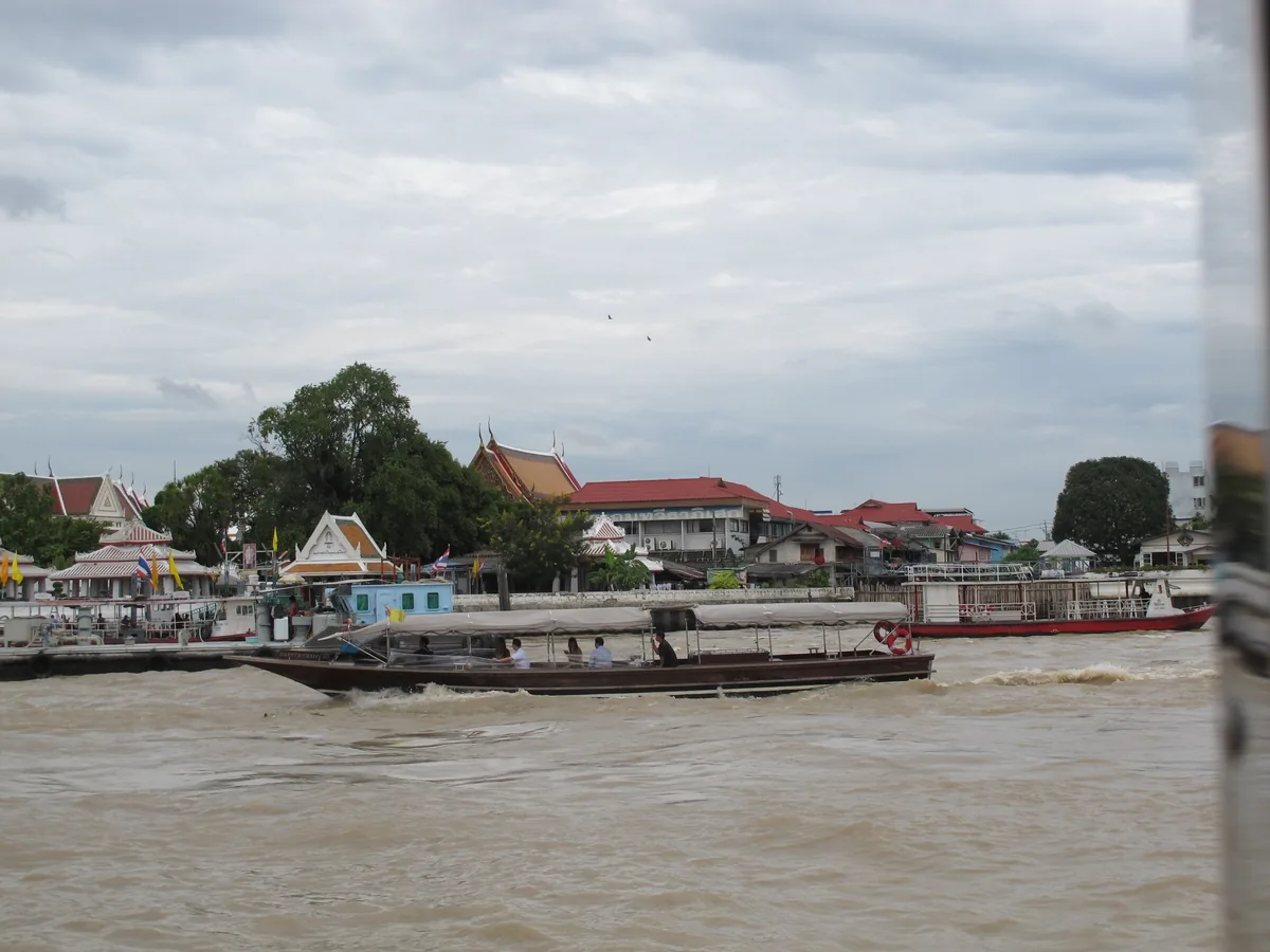 A long-tail boat carries passengers along the muddy Chao Phraya River past temple roofs in Bangkok.