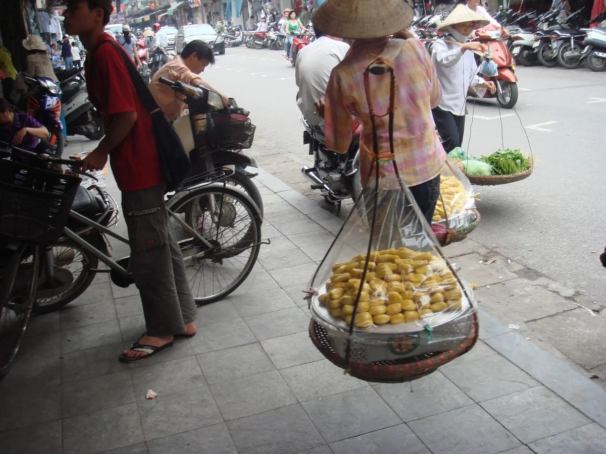 A conical hat street vendor carries baskets of jackfruit through a busy sidewalk in Hanoi, Vietnam.