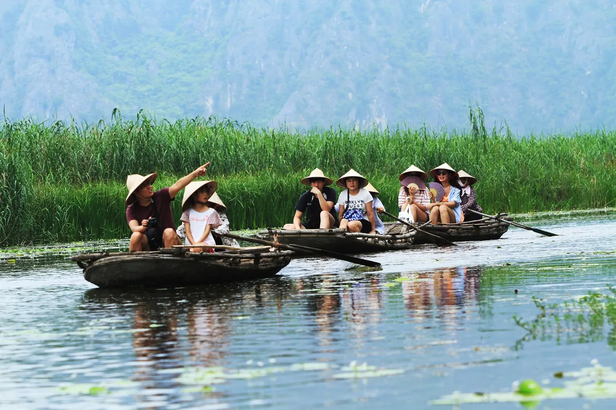 Tourists in conical hats ride small wooden boats through reeds beneath limestone mountains in Ninh Binh.