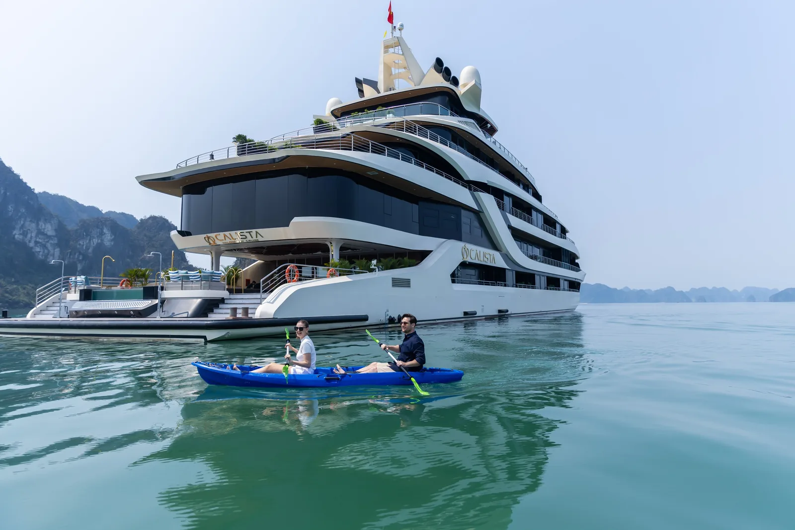 A couple kayaks past the luxury Calista cruise ship anchored on Halong Bay's calm waters.
