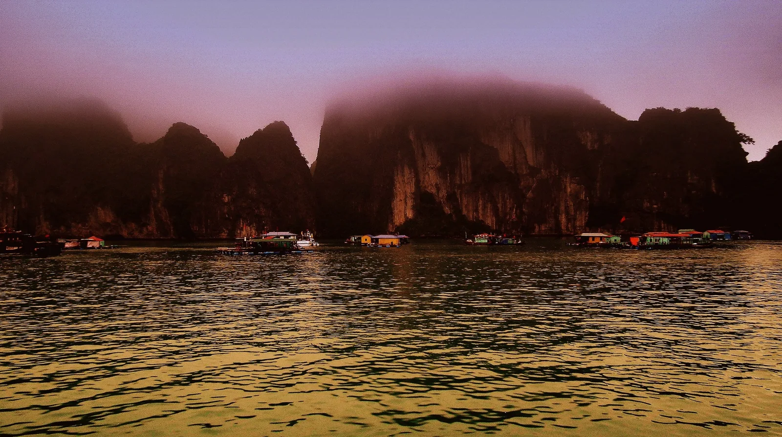 A floating fishing village nestles beneath mist-shrouded limestone karsts in Ha Long Bay at dusk.