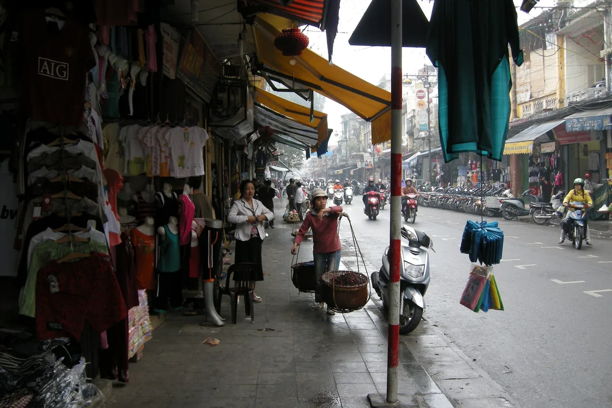 A street vendor carries a shoulder pole past clothing stalls and parked motorbikes on a busy Hanoi sidewalk.