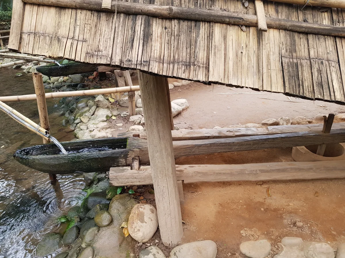 Bamboo water pipes carry a stream into carved wooden troughs beside a thatched hut at an open-air ethnology museum in Hanoi.
