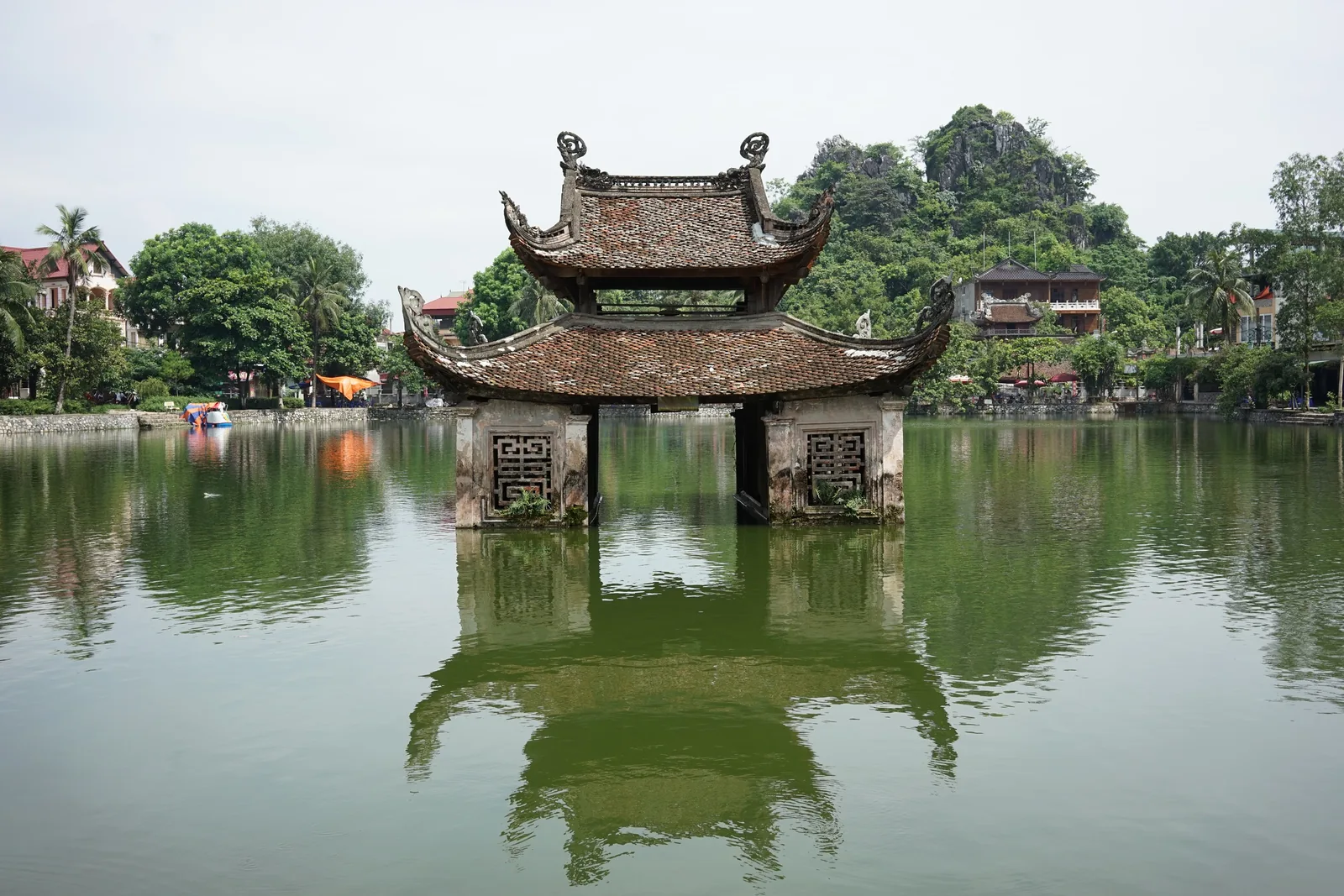 A traditional stilted pavilion with tiled curved roofs stands over a green pond near Hanoi, with limestone hills in the.
