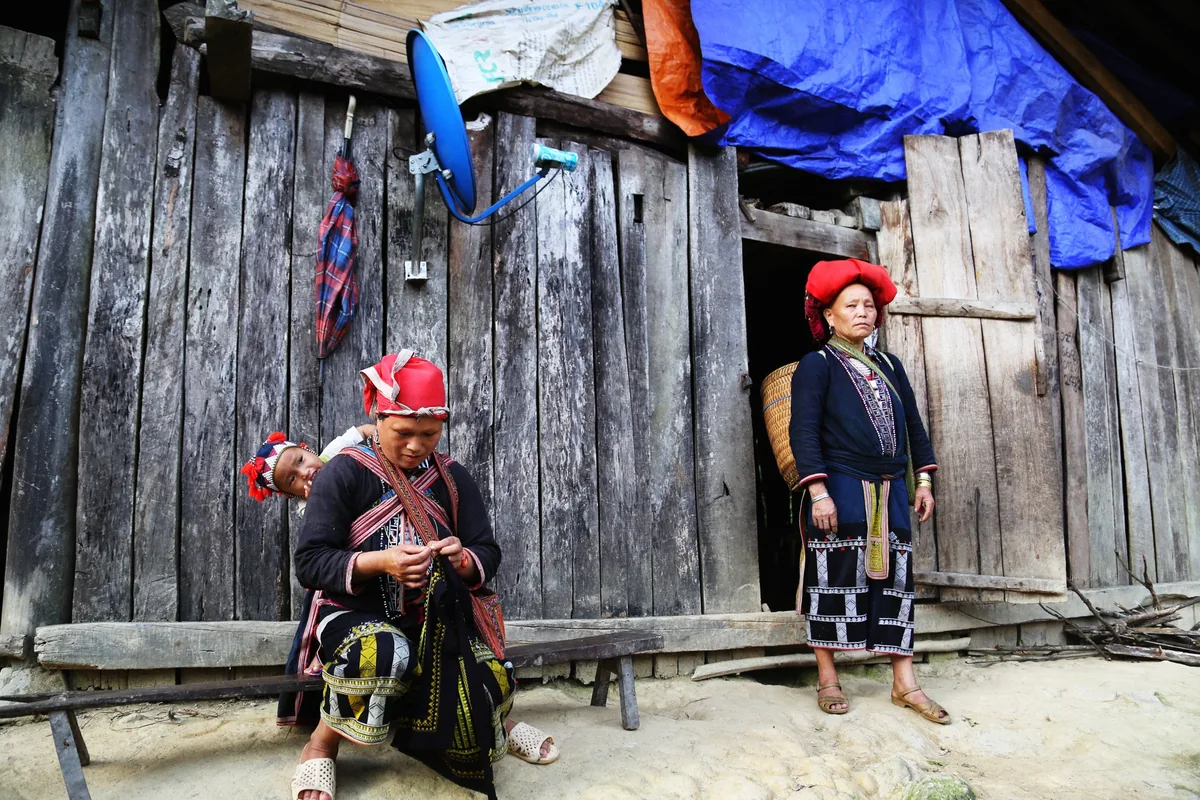 Red Dao women in traditional embroidered clothing and turbans stand outside a wooden house near Sapa
